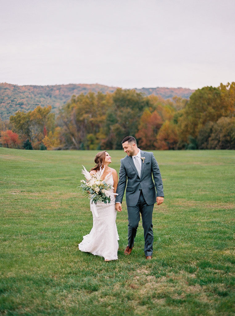 A couple walks together through the northern Virginia countryside, holding hands and smiling at each other. Photographed by northern Virginia wedding photographer Kim Branagan.