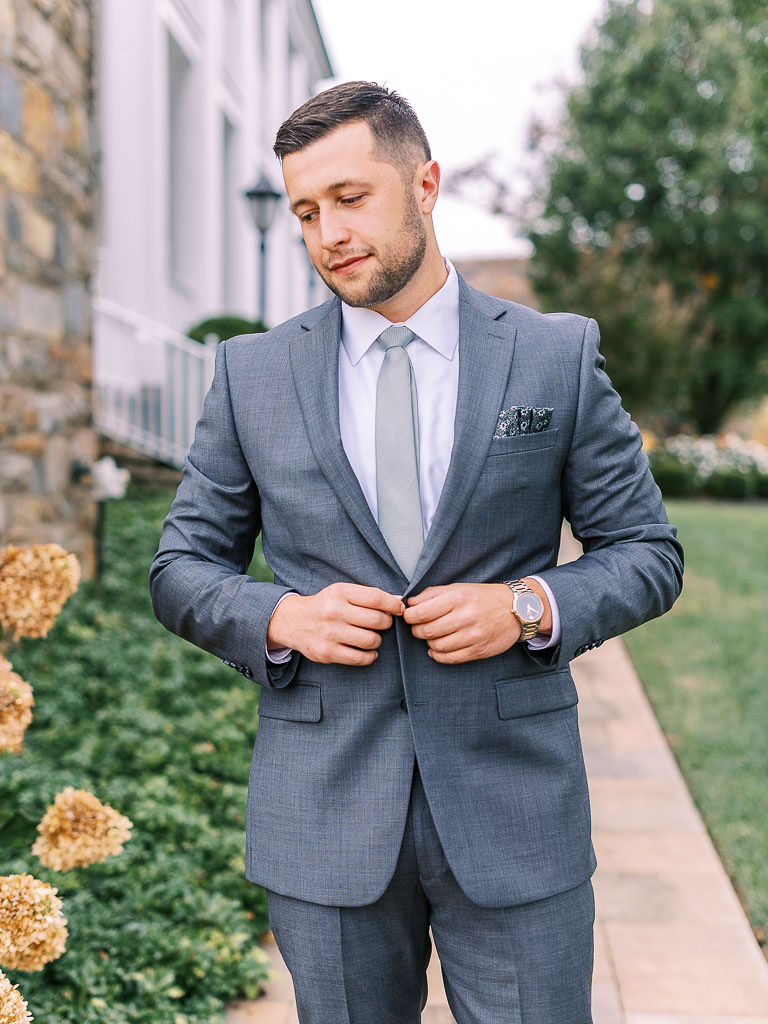A man in a suit buttoning his suitcoat standing next to some flowers on his wedding day