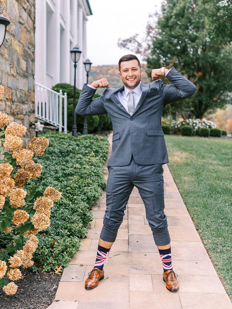 A man jokingly flexing his arm muscles on his wedding day. He is wearing a suit, with socks with the British flag on them.