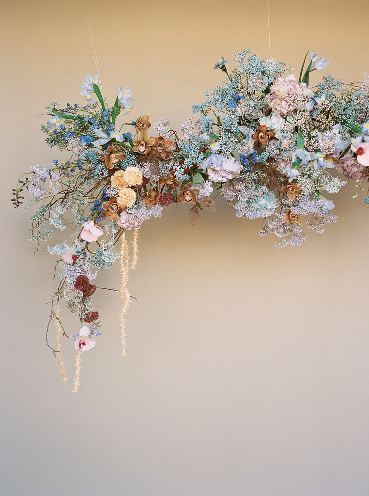 A vibrant hanging floral arch installation against a golden-white wall. The flowers in the arrangement are pastel mauve, pink, orange, and blue with lots of greenery.