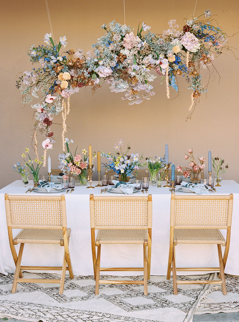 During a branding editorial photo shoot, a boho-styled table with wicker chairs, colorful candles, and spring wildflower bouquets is on a woven rug, with a hanging floral installation that matches the table centerpieces hanging above.