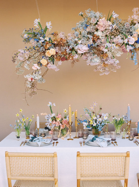 A vibrant, spring-themed floral arrangement hangs about a boho-styled table. The table has individual place settings, colorful candles, and wildflower bouquets on it. Designed by Lavenders Flowers in Santa Ana, CA and photographed by California branding photographer Kim Branagan