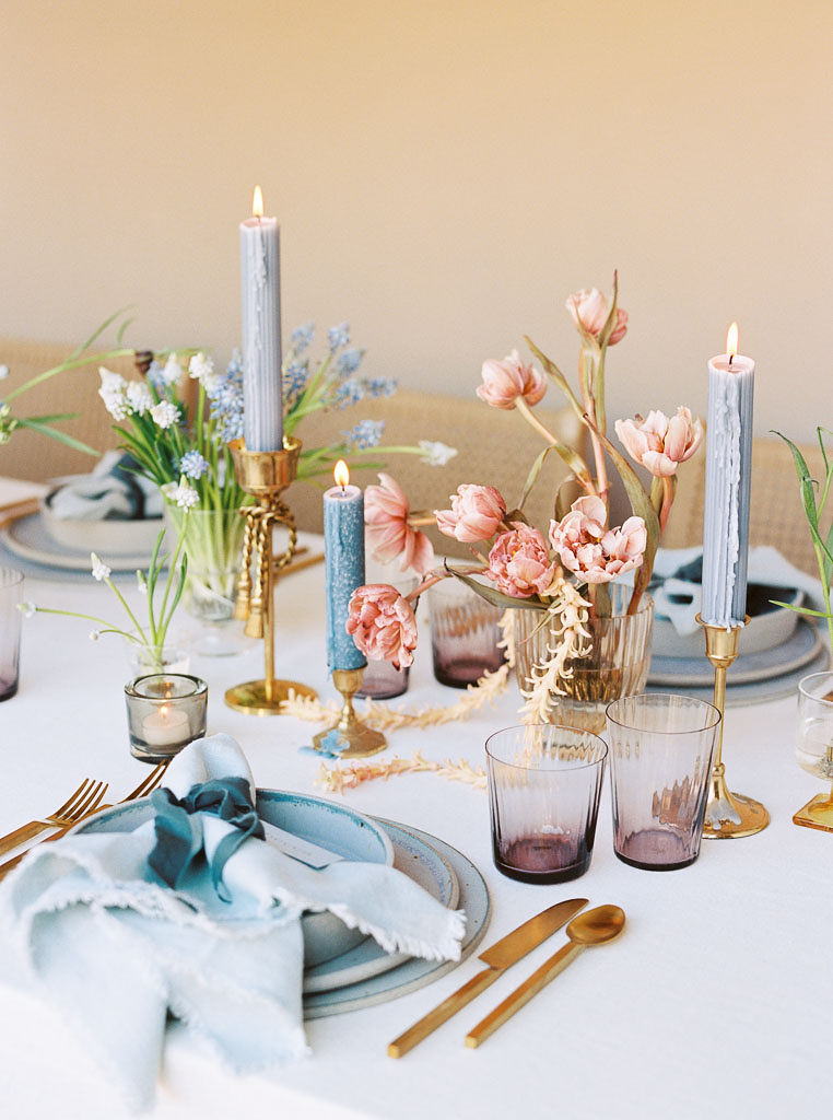 A styled table setup designed with a spring theme. Tall, pale blue candles and pink wildflowers line the center of the table. Individual place settings are set on the table with ceramic plates and pale blue napkins