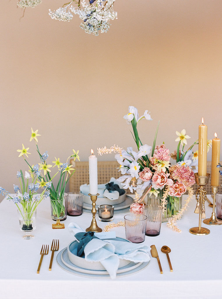 A table setting with several golden and white candles, several wildflower bouquets, copper silverware, and pale blue napkins