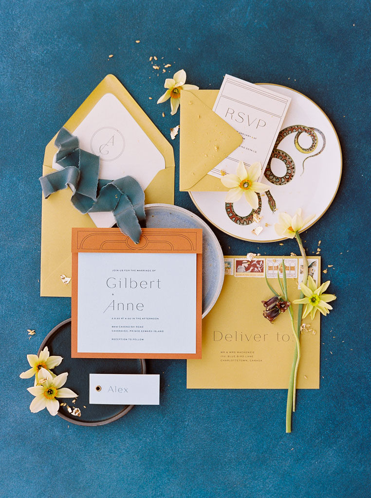 Party invitations in mustard yellow and burnt orange colors lay on unique pottery plates with individual wildflowers and ribbons arranged over them. Everything is laying on a deep blue backdrop. Photographed by California branding photographer Kim Branagan for a branding editorial photo shoot with Lavenders Flowers (Santa Ana, CA.)