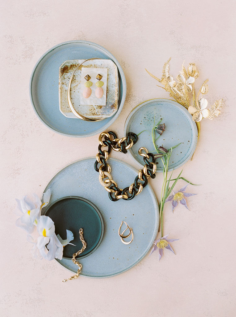 Pale blue pottery plates with gold jewelry on them and single wildflowers laying by them, too. Everything is arranged on a pale mauve backdrop.