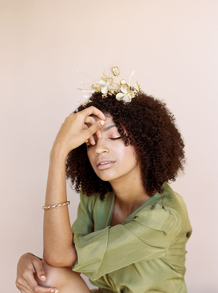 A model at a branding editorial photo shoot puts her hand on her face as she closes her eyes. She's wearing an olive green dress and a golden hair piece