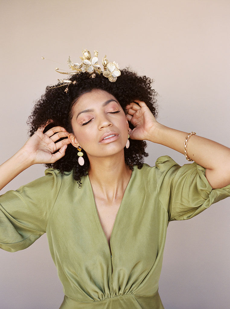 A mode at a branding editorial photo shoot closes her eyes as she pushes her hair back from her face. She is wearing a golden floral hair piece and golden jewelry. Shot by editorial photographer Kim Branagan in Santa Ana, CA