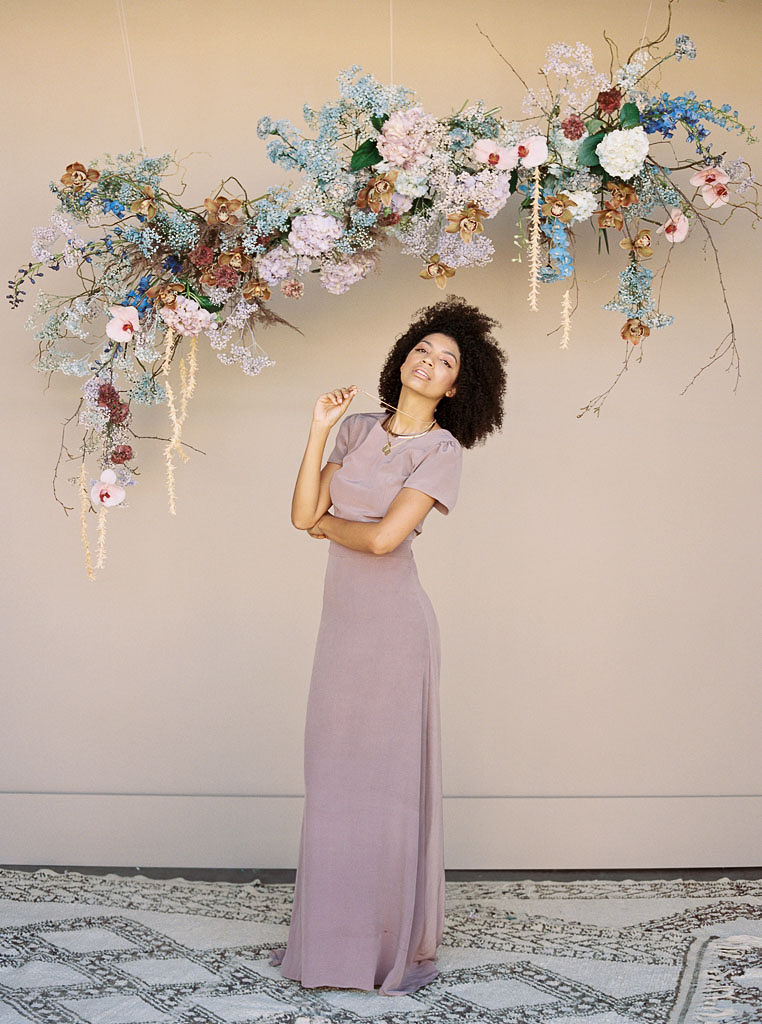 A model at a branding editorial photo shoot stands in a mauve colored maxi dress on a woven rug in front of a colorful, spring-theme floral arrangement hanging on a wall