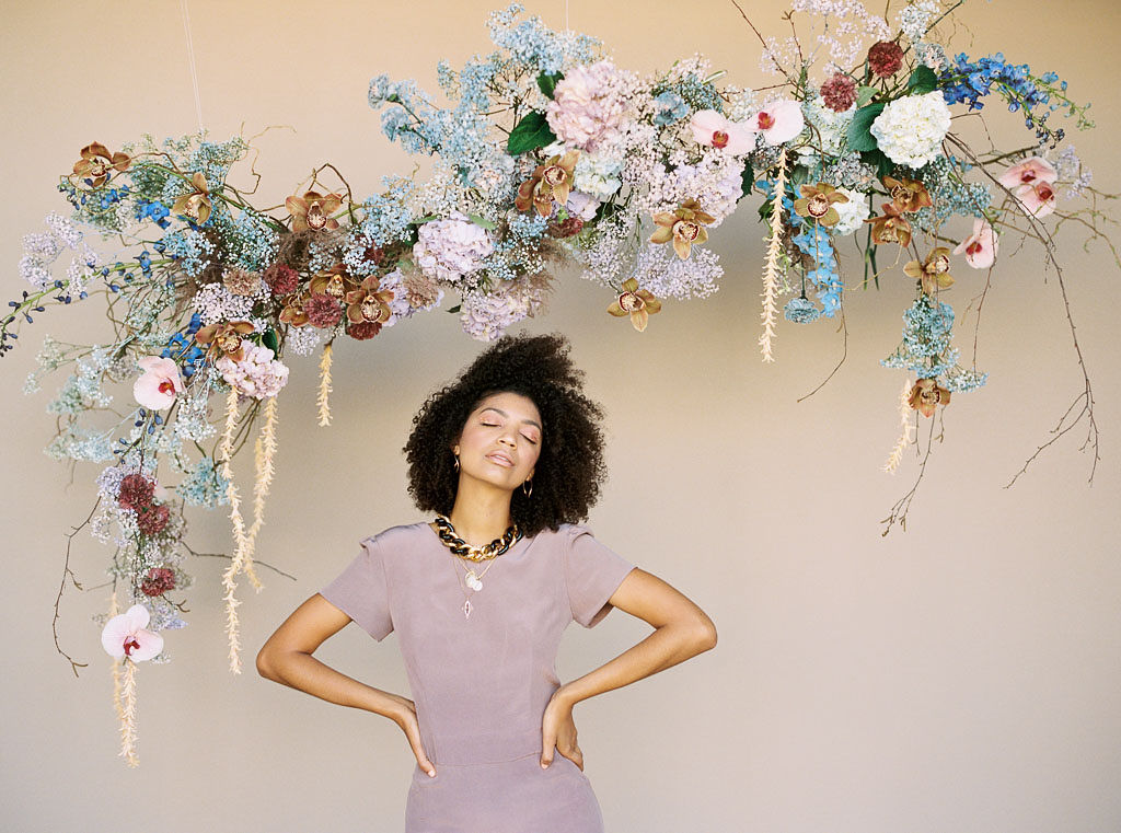 A woman at a branding editorial photo shoot wearing a lavender colored dress and chunky gold necklaces stands with her hands on her hips and her eyes closed in front of pastel-colored, spring-themed floral arch hanging on the wall behind her. Photographed by California brand photographer Kim Branagan