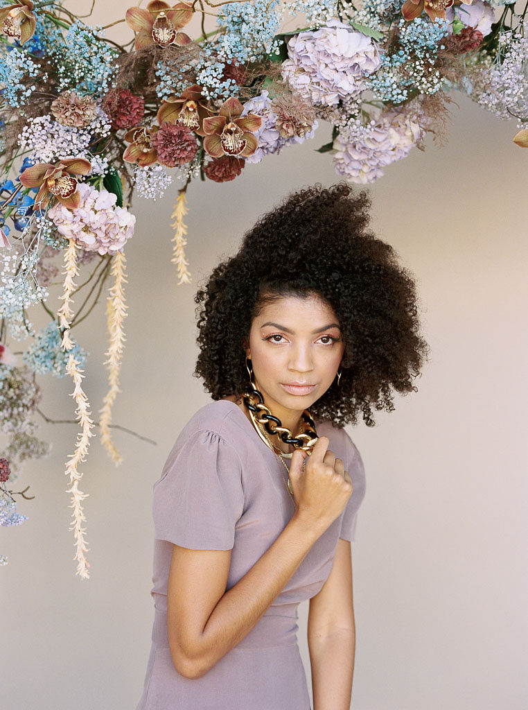A woman at a branding editorial photo shoot touches a gold chain necklace she is wearing and stares straight ahead. She is standing in front a hanging floral arrangement comprised of mauve, blue, and terra cotta brown flowers
