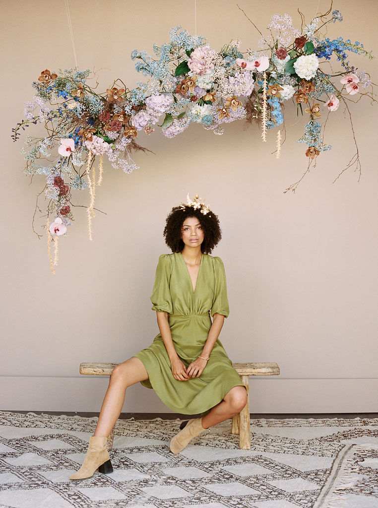 A model at a branding editorial photo shoot with a floral hairpiece sitting on a wooden bench and woven rug under a colorful hanging floral arrangement designed by Lavenders Flowers for a branding editorial photo shoot. Photographed by California branding photographer Kim Branagan
