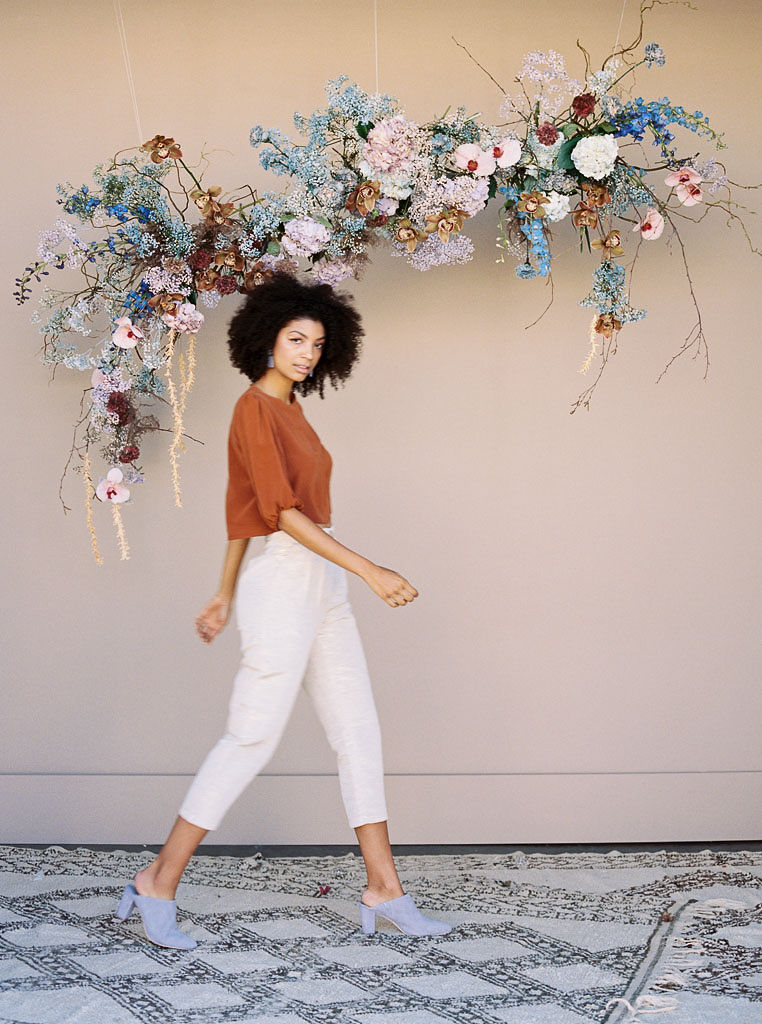 A woman wearing a casual outfit of a burnt orange blouse, white capri pants, and pastel blue blogs walks on a woven rug in front of a floral installment comprised of pastel blue, mauve, pink, and burnt orange flowers. Photographed by southern California editorial photographer Kim Branagan.