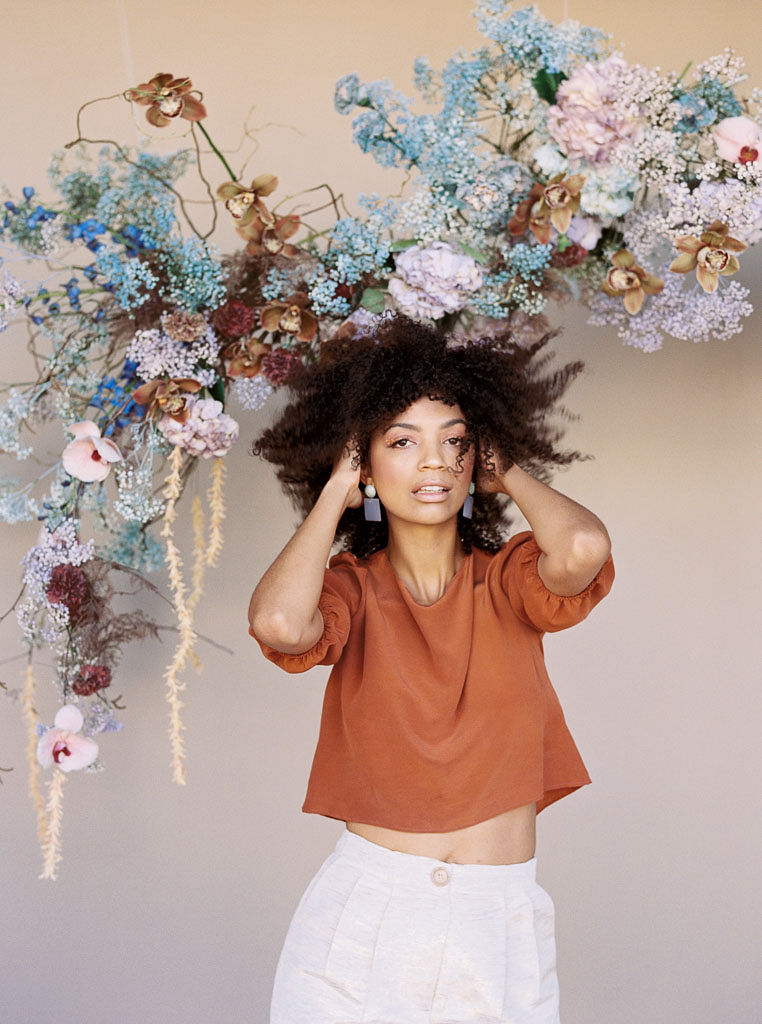 A woman at a branding editorial photo shoot fluffs her hair with hands. She is wearing a burnt orange blouse and pale blue earrings. She is standing in front of a vibrant, spring-themed floral arch that matches her outfit. Photographed for Lavenders Flowers in Santa Ana, California for a branding editorial photo shoot by Kim Branagan