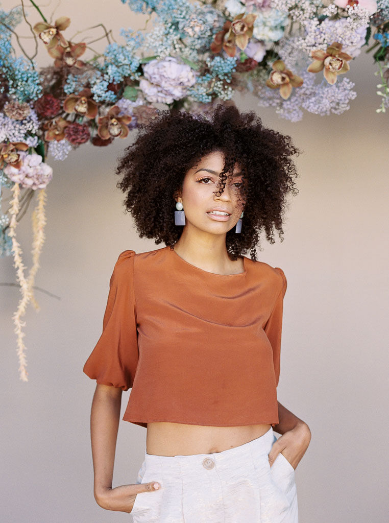 A model stares into the camera while standing in front of blue, red, and mauve hanging floral installation. She is wearing a burnt orange cropped blouse, and her hands are in the pockets of her white linen pants