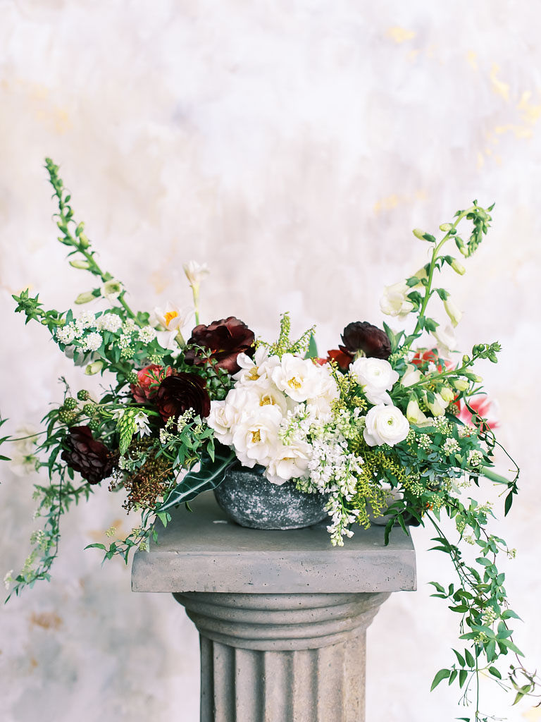 A floral arrangement with white and maroon flowers and lots of greenery against a marble wall