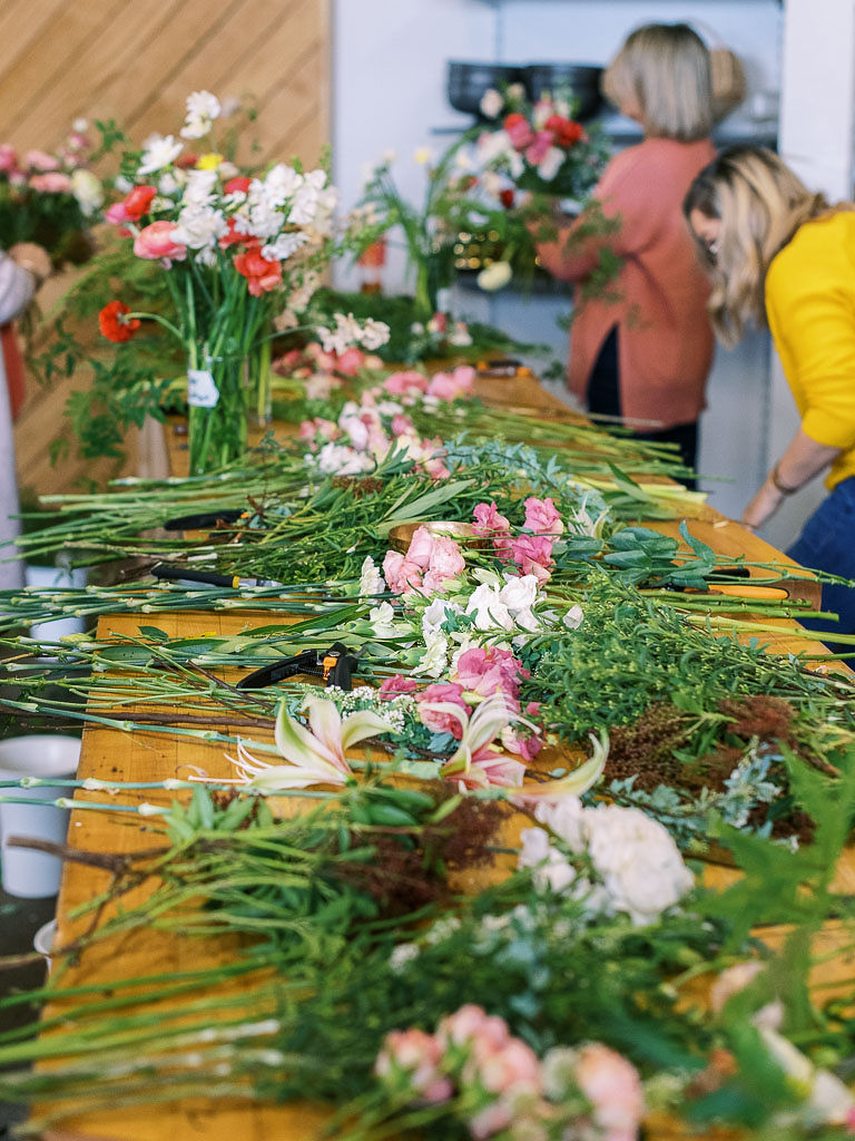 Flowers with long stems laid out across a long wooden table as floral workshop attendees work on their bouquets. Photographed by branding and event photographer Kim Branagan