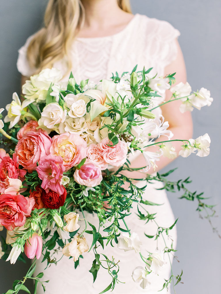 A woman wearing a white dress holds a lush, cascading bouquet of pink, red, peach, and ivory roses