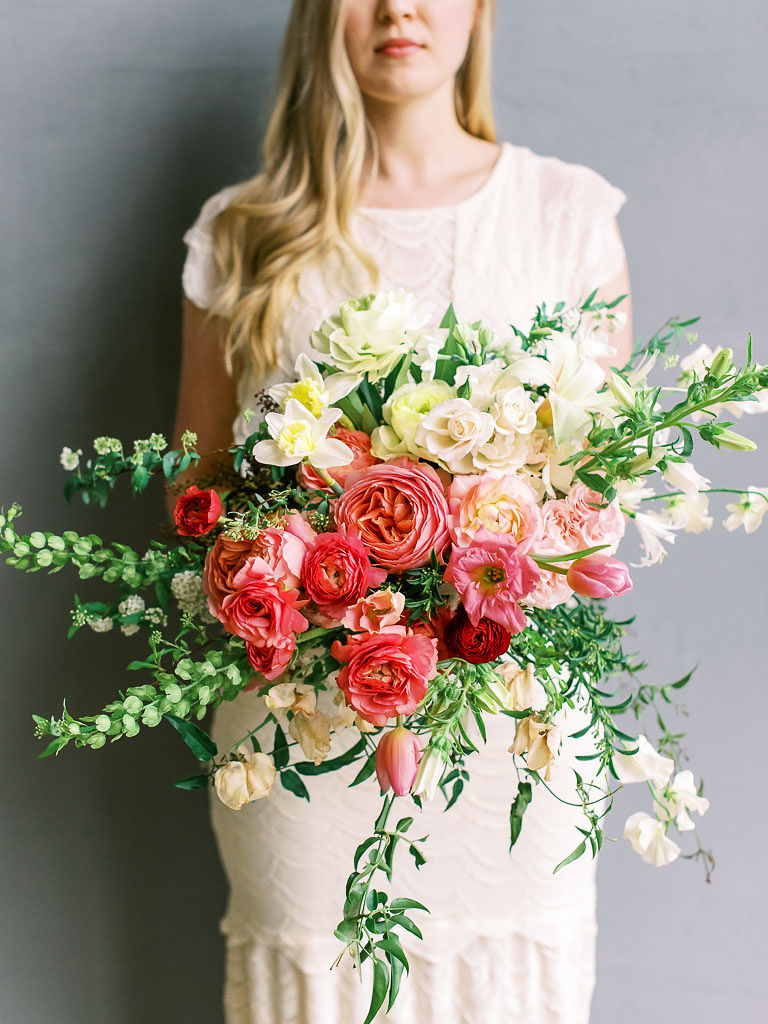 A woman wearing a white dress holds a blush, pink, and cream floral bouquet against a minimal gray backdrop. Photographed by northern Virginia floral photographer Kim Branagan