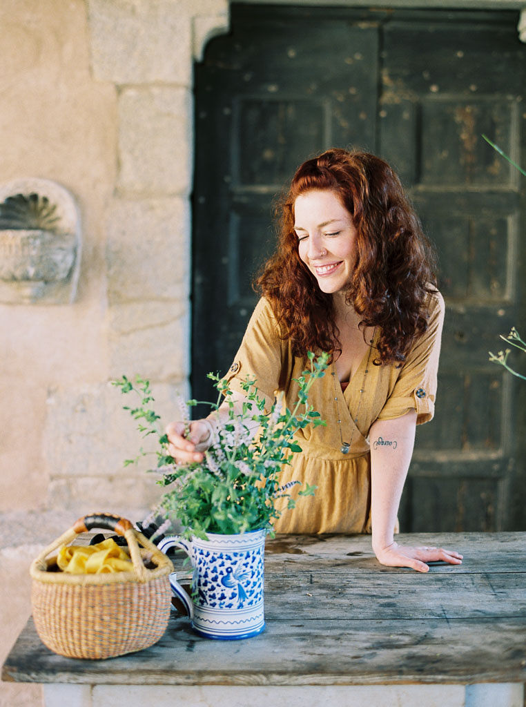 A woman arranging flowers for a floral design workshop. She is working on a wooden table with a stone wall behind her. Clarity Retreat - A Floral Workshop by Ponderosa & Thyme | Florence, Italy. Photographed by Florence brand and editorial photographer Kim Branagan.