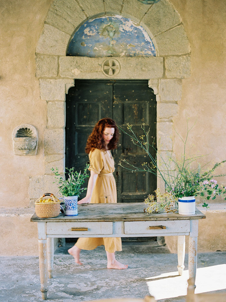 A woman walking behind a wooden table with flowers in vases on it. There is a stone wall behind the woman. Clarity Retreat - A Floral Workshop by Ponderosa & Thyme | Florence, Italy. Photographed by Florence brand and editorial photographer Kim Branagan.