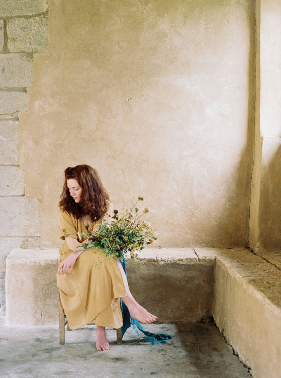 Katie Davis of Ponderosa and Thyme in a yellow dress looks down at the ground as she sits on a chair in front of a textured stone wall, holding a floral bouquet.
