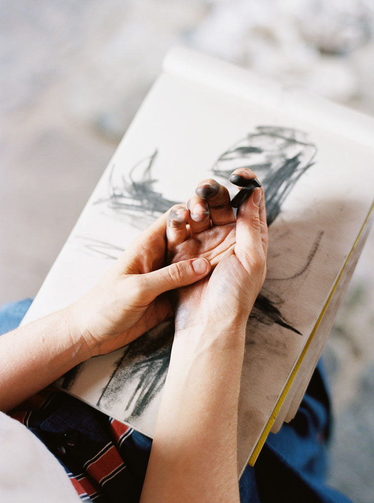 Close up shot of a person's hands as they work on a charcoal drawing. The person's hands are tinged black with charcoal.