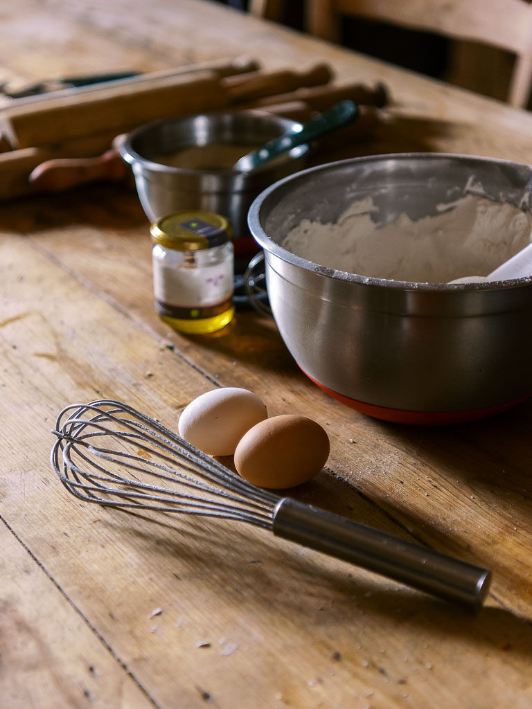 Flour, eggs, honey, a whisk, and a rolling pin on a wooden table.