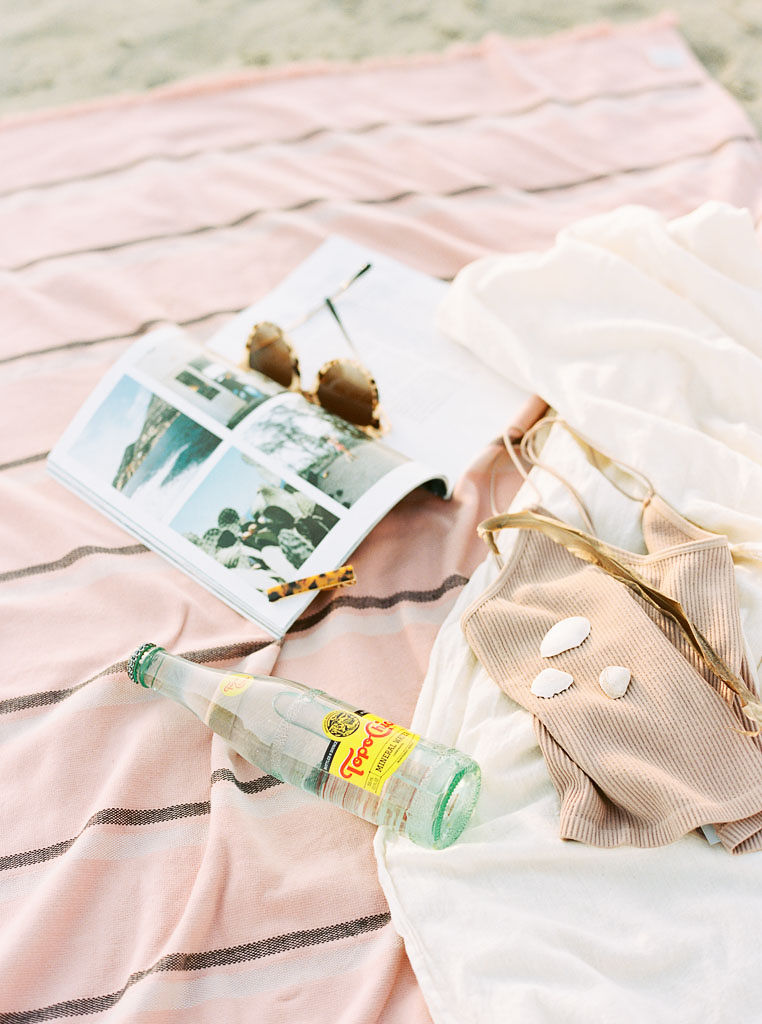 A pink, striped beach blanket with a magazine, Top Chico bottle, and seashells on it. Destination Photoshoot for a Lifestyle Editorial on the Beach in San Diego, CA by San Diego lifestyle editorial photographer Kim Branagan