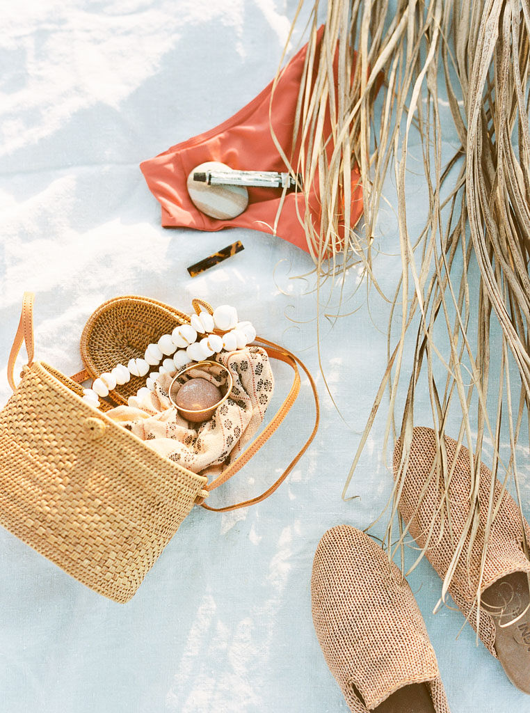 A wicker purse, woven slip on shoes, beauty products, a swim suit and palm branch on a white beach blanket. Destination Photoshoot for a Lifestyle Editorial on the Beach in San Diego, CA by San Diego lifestyle editorial photographer Kim Branagan