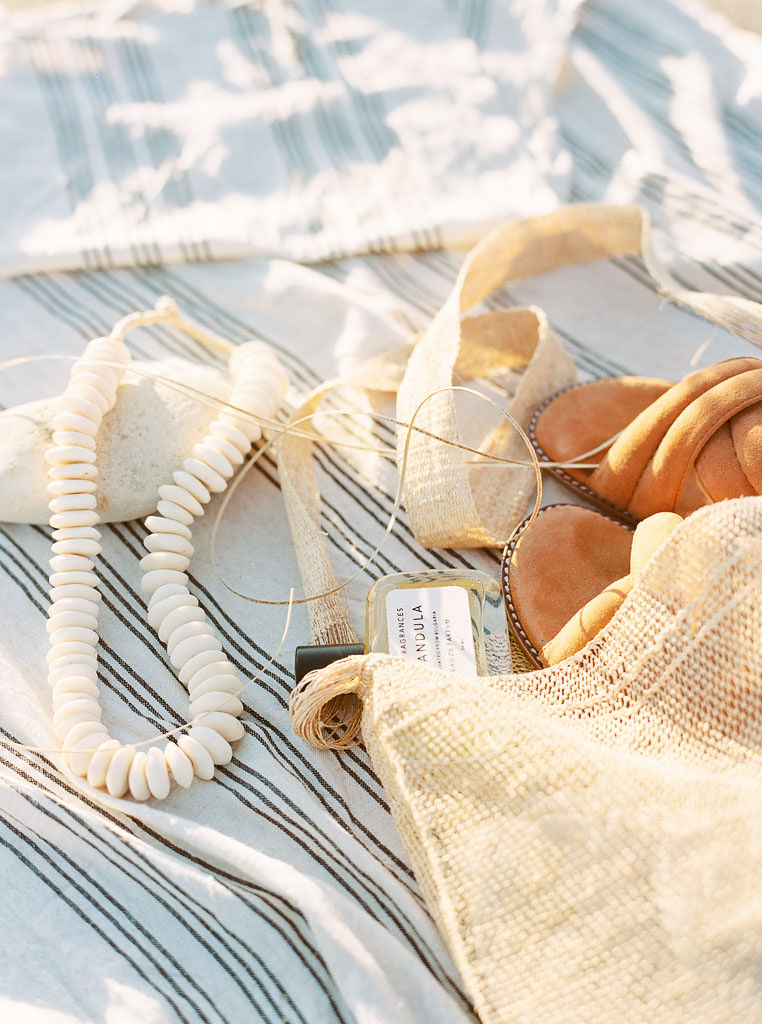 Close up shot of a shell necklace, a woven beach tote, and peach sandals on a white and black striped beach blanket. Destination Photoshoot for a Lifestyle Editorial on the Beach in San Diego, CA by San Diego lifestyle editorial photographer Kim Branagan