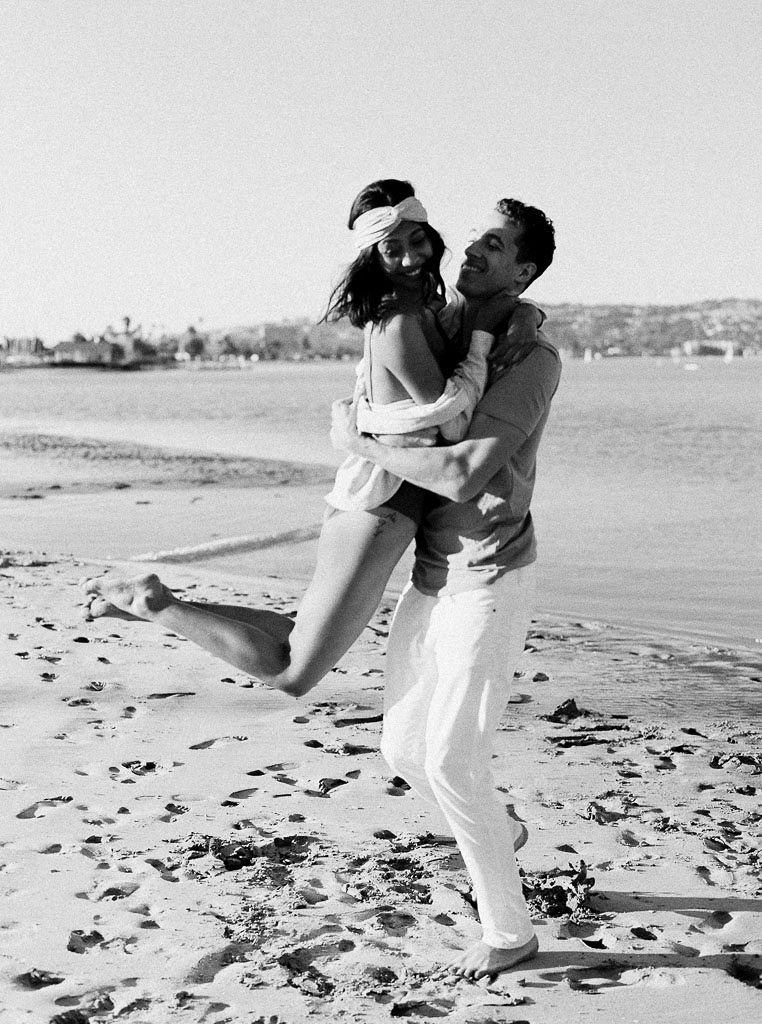 A man lifting up a woman and twirling her around as they stand on the beach. Destination Photoshoot for a Lifestyle Editorial on the Beach in San Diego, CA by San Diego lifestyle editorial photographer Kim Branagan