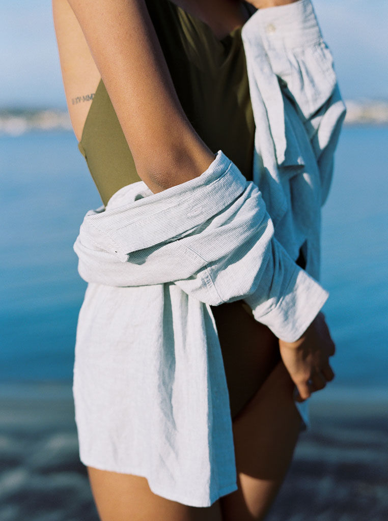 Close up shot of a woman wearing a sleek sporty green one piece swim suit and linen, long sleeve shirt with her arms crossed over her. Destination Photoshoot for a Lifestyle Editorial on the Beach in San Diego, CA by San Diego lifestyle editorial photographer Kim Branagan
