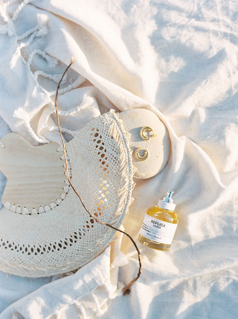 A hand-held fan,gold earrings, and perfume bottle on a white linen beach blanket. Destination Photoshoot for a Lifestyle Editorial on the Beach in San Diego, CA by San Diego lifestyle editorial photographer Kim Branagan