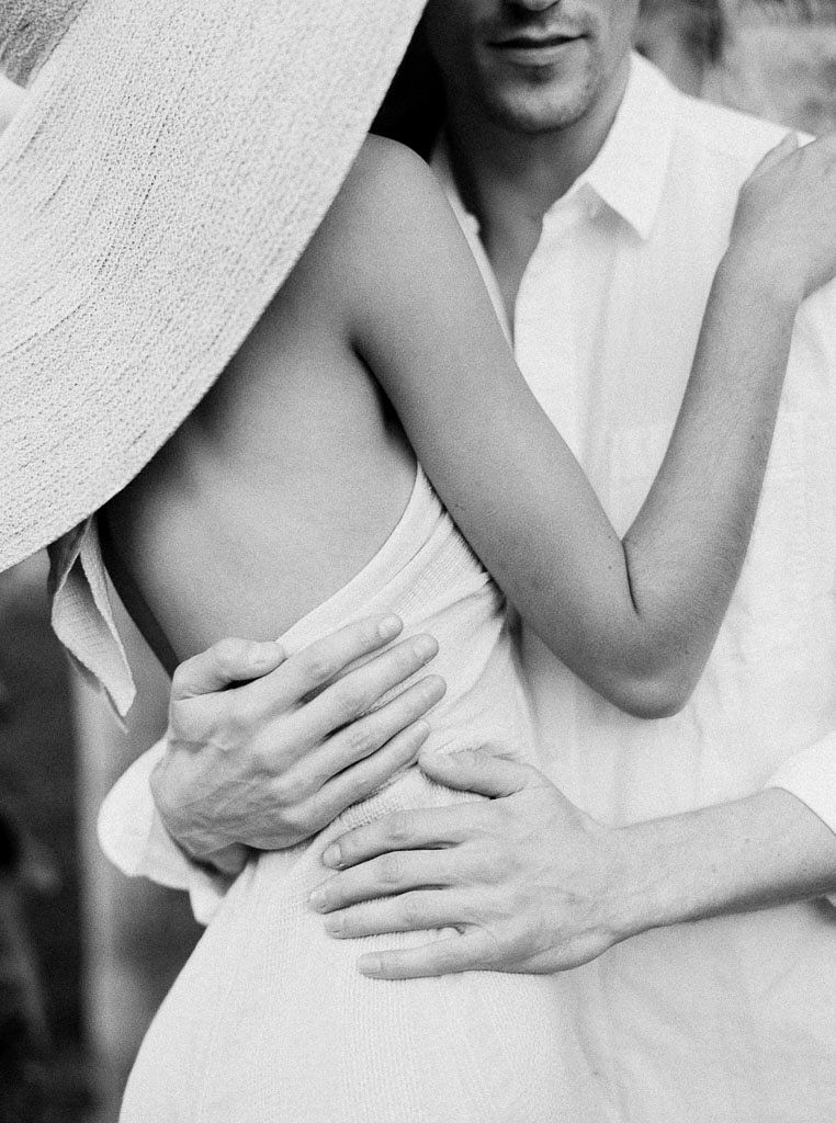 Close up shot of a woman wearing a white dress and wide-brimmed sun hat embracing a man wearing a white linen button-down shirt. Destination Photoshoot for a Lifestyle Editorial on the Beach in San Diego, CA by San Diego lifestyle editorial photographer Kim Branagan