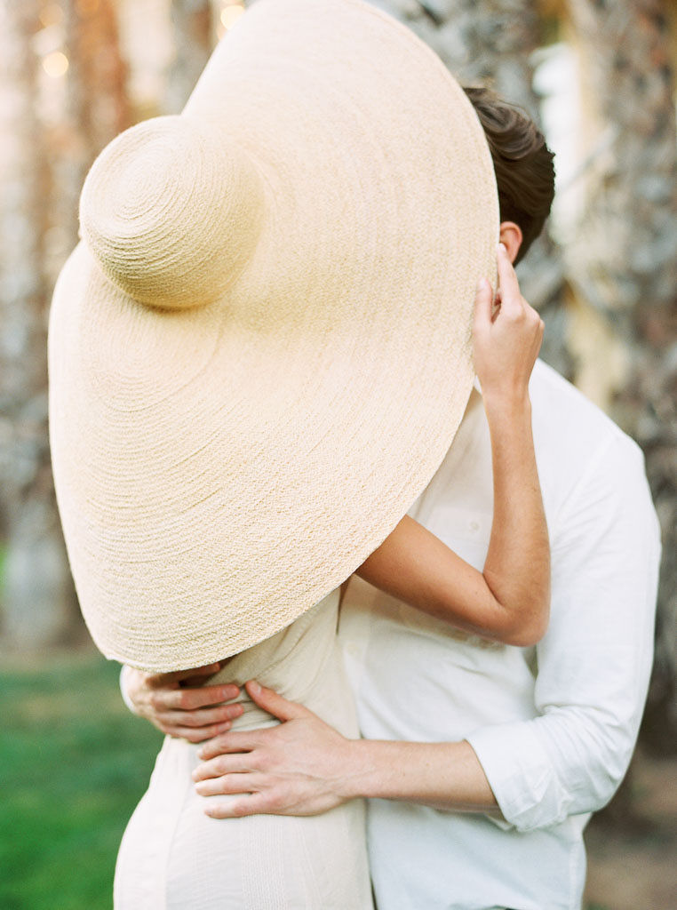 A couple embraces, and the woman pulls the wide brim of her large sun hat over the faces to hide from the camera. Destination Photoshoot for a Lifestyle Editorial on the Beach in San Diego, CA by San Diego lifestyle editorial photographer Kim Branagan