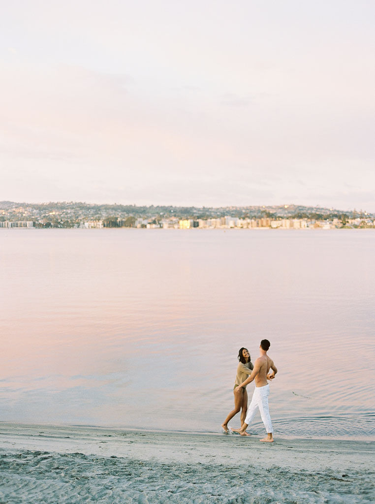 A couple plays around in the water at the shoreline as the sunset reflects pink light over the water. Destination Photoshoot for a Lifestyle Editorial on the Beach in San Diego, CA by San Diego lifestyle editorial photographer Kim Branagan