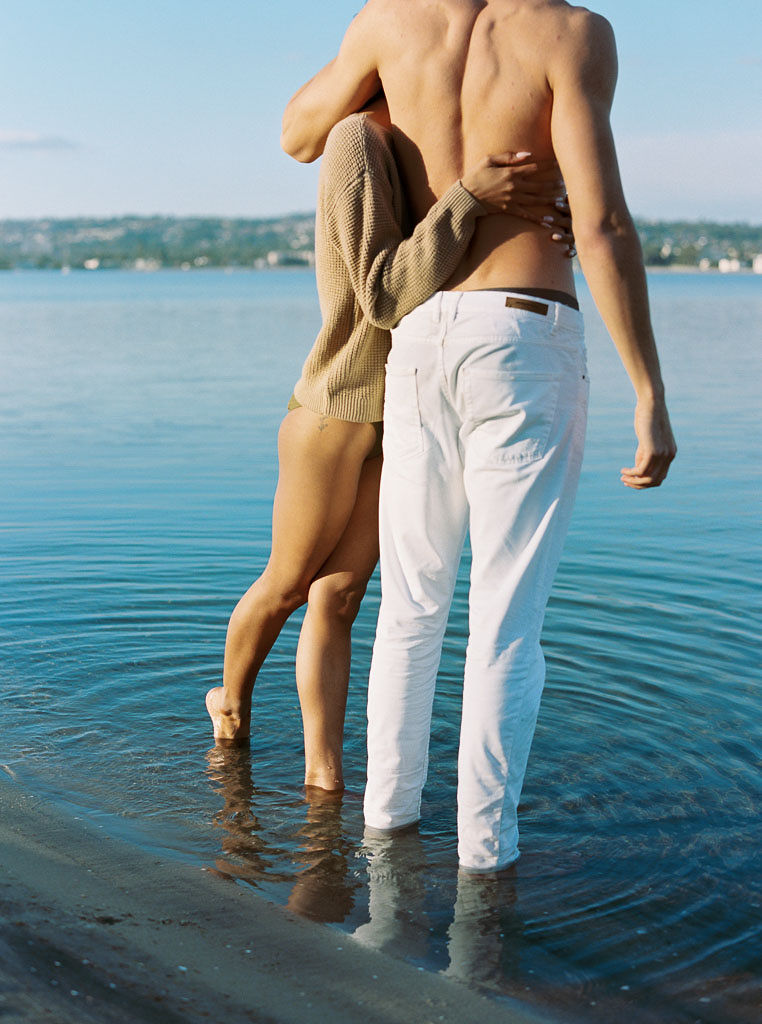 A woman wear a sweater over her one piece swim suit hugging a man standing next to her, who is shirtless and wearing white denim jeans. They are standing at the shoreline of the beach.