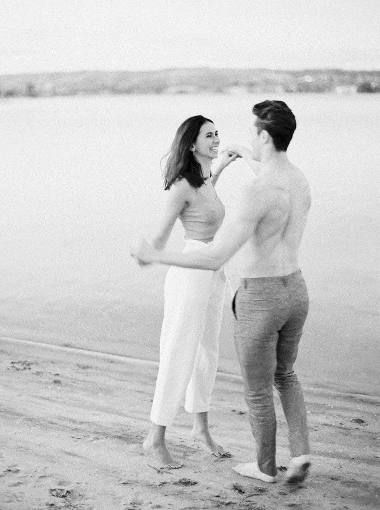 A couple smiles and laughs as they dance together on the beach right in front of the water. Destination Photoshoot for a Lifestyle Editorial on the Beach in San Diego, CA by San Diego lifestyle editorial photographer Kim Branagan
