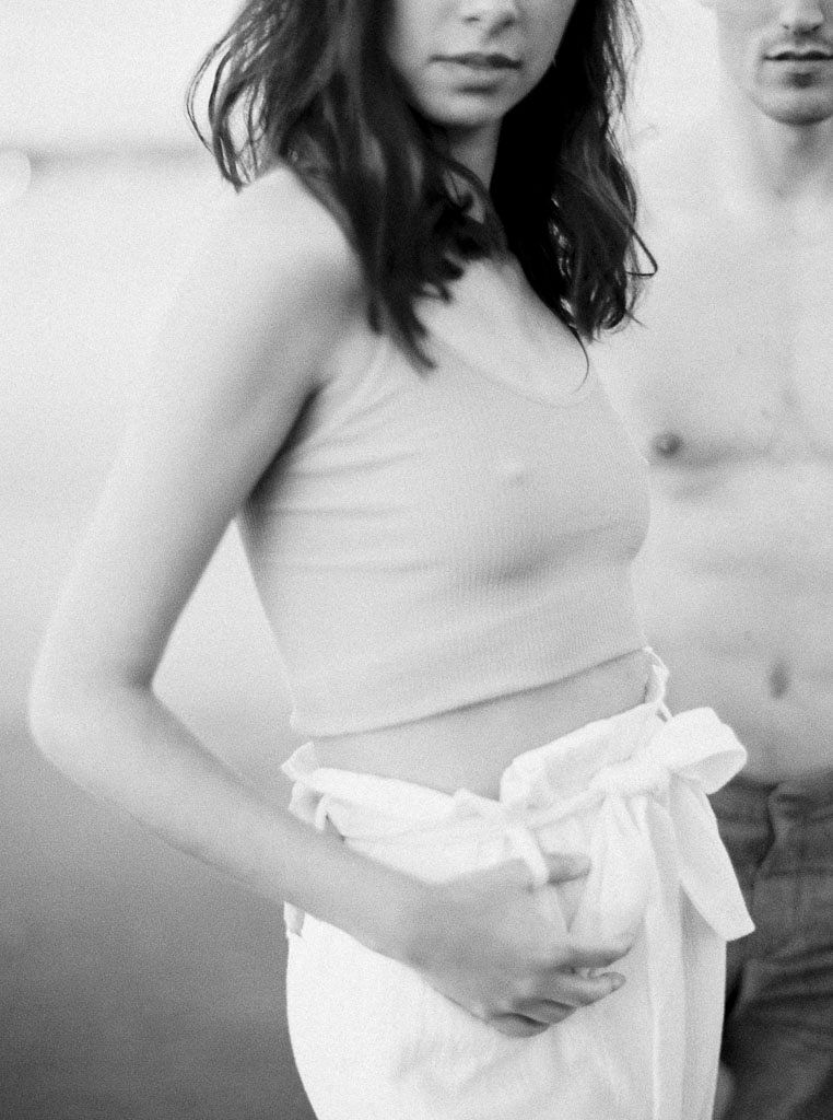 Close up shot of a woman with dark brown hair wearing a cropped tank top and white linen pants. A shirtless man is standing behind her. Destination Photoshoot for a Lifestyle Editorial on the Beach in San Diego, CA by San Diego lifestyle editorial photographer Kim Branagan