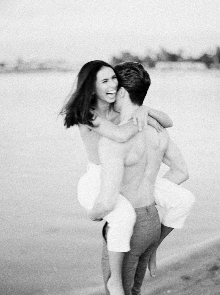 A man picks up a woman and carries her. The woman is laughing. Destination Photoshoot for a Lifestyle Editorial on the Beach in San Diego, CA by San Diego lifestyle editorial photographer Kim Branagan