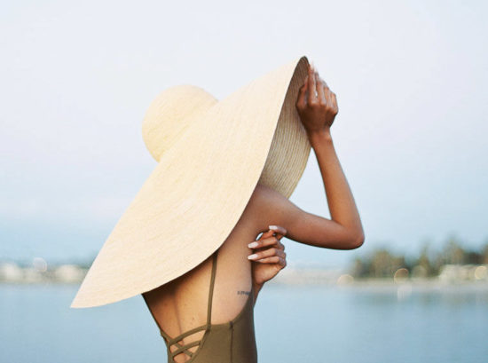 A woman wearing an olive green one piece swimsuit and large, wide-brimmed woven hat stands at the shoreline at the beach, with her back to the camera. Destination Photoshoot for a Lifestyle Editorial on the Beach in San Diego, CA by San Diego lifestyle editorial photographer Kim Branagan